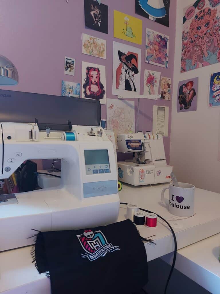 photo d'un bureau blanc, brodeuse brother au premier plan avec bobines de fil à broder et broderie monster High. au fond, surjeteuse, tasse à café I love toulouse. Mur du fond violet pastel et illustrations de divers artistes