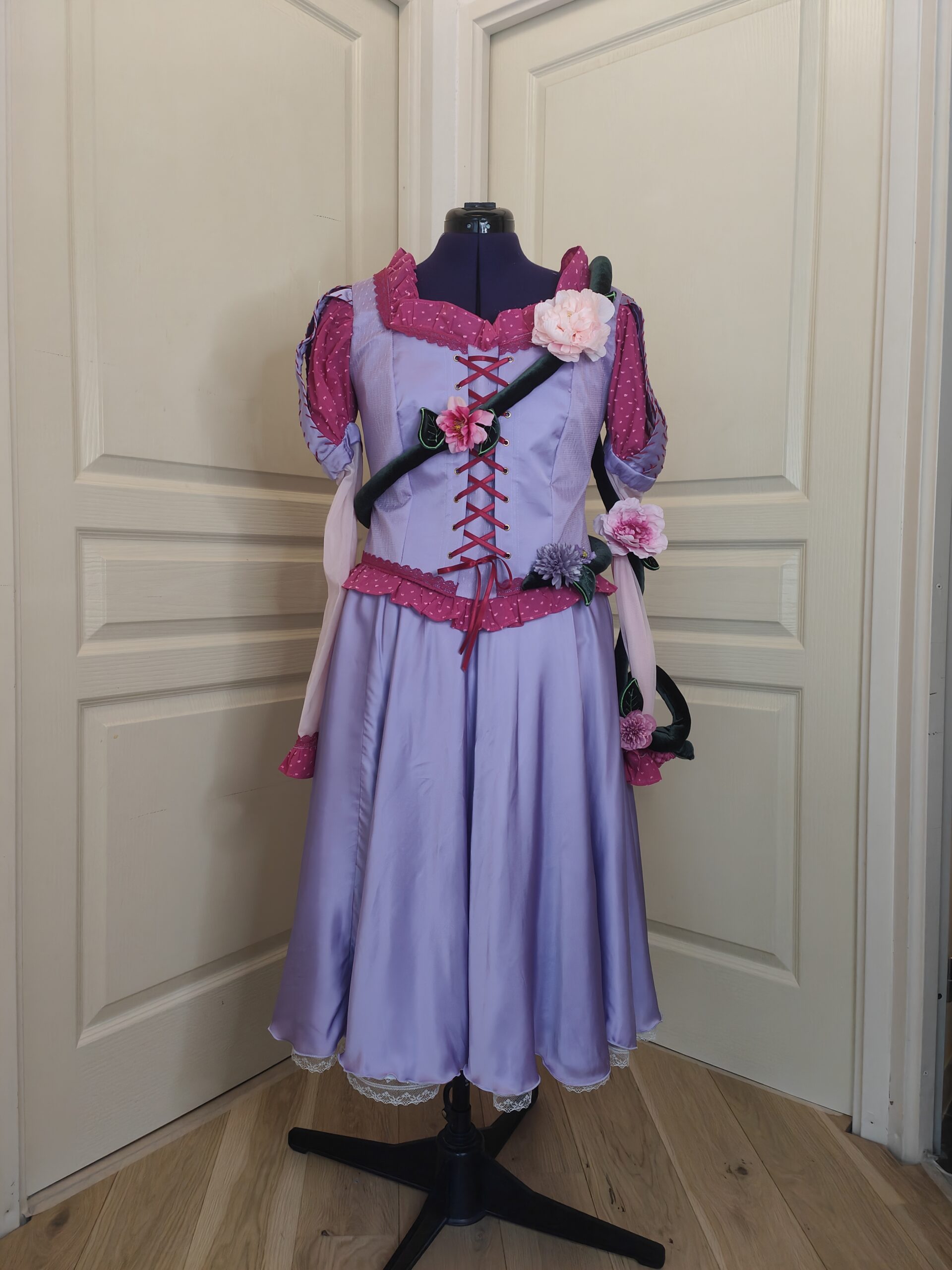 Costume de Raiponce Pirates et Princesses. Robe de couleur lavande avec détails et laçages rose vifs. Liane avec fleur et feuilles brodées enroulée autour du buste et bras gauche.